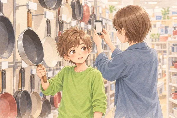 台所用品の売り場。壁にフライパンがずらりと吊るされ、若草色の長袖のかえでが、1枚を片手で持ち上げて、少し驚いた顔で笑っている。右の手前で、灰みの青の上着のみはるさんが、スマホで札を撮っている